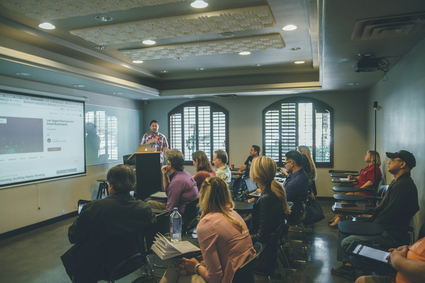 Teachers seated at tables in a bright room listening to a presenter